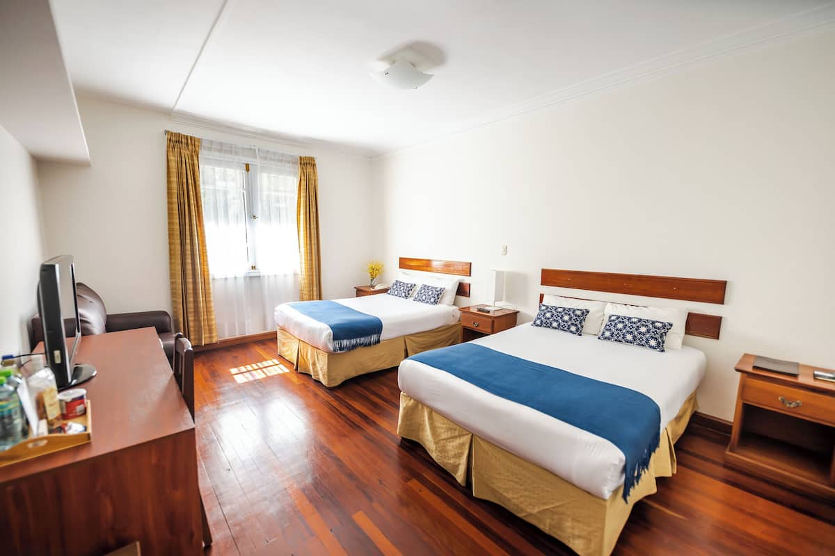 A hotel room with two beds, wooden flooring, a desk with a computer, and a window with curtains.