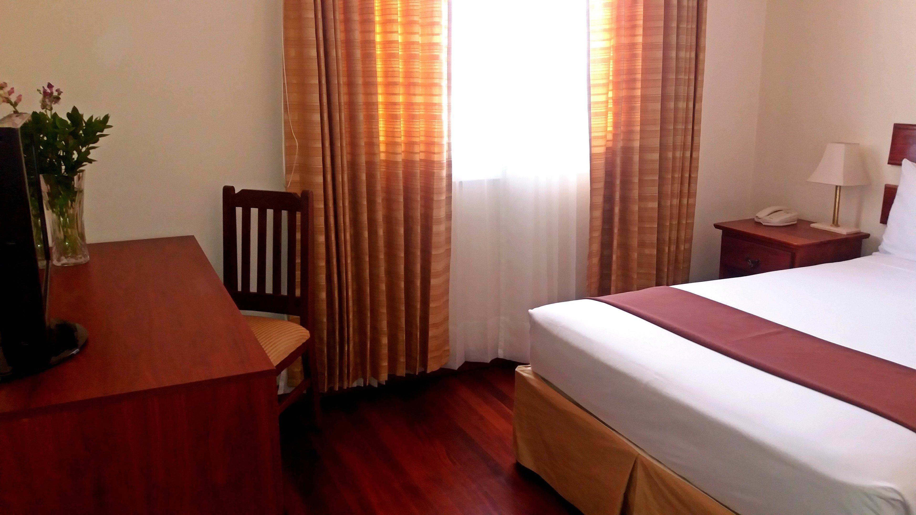 A hotel room with a wooden desk, a chair, a bed with a red bedspread, a nightstand with a lamp, and a telephone.