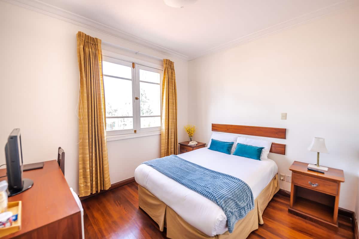 A hotel room with a bed, bedside table, lamp, and a desk with a computer.