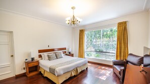Quarto com piso de madeira, cama com cabeceira, mesa de cabeceira, janela com cortinas e uma poltrona de couro marrom.
