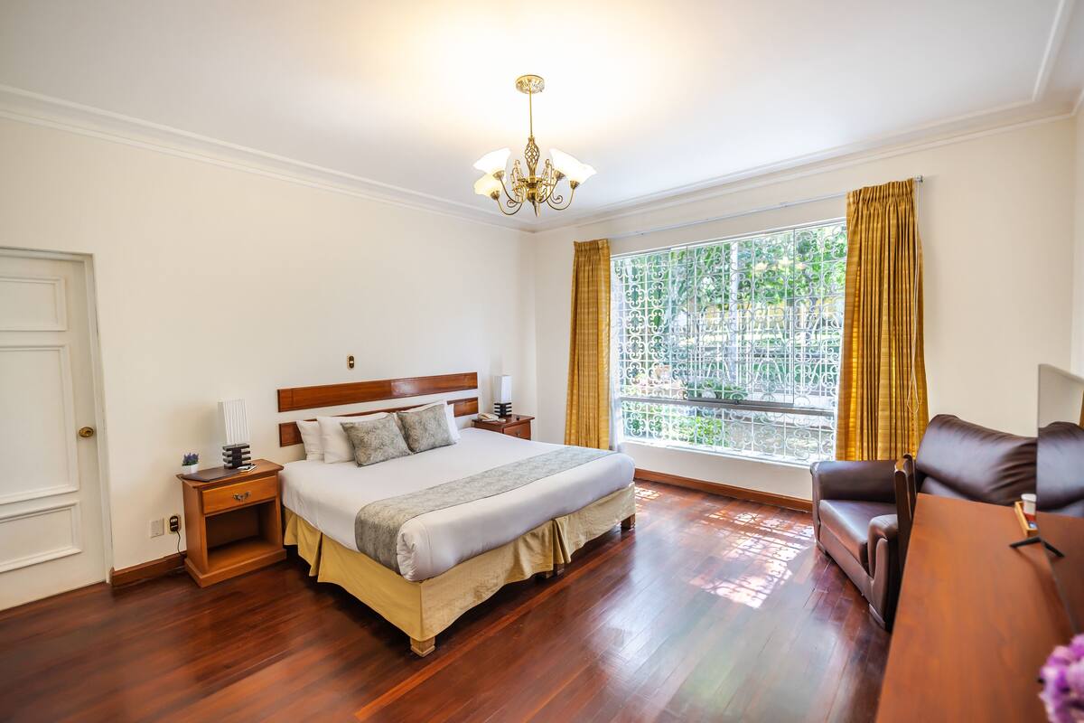 A bedroom with a wooden floor, a bed with a headboard, a nightstand, a window with curtains, and a brown leather chair.