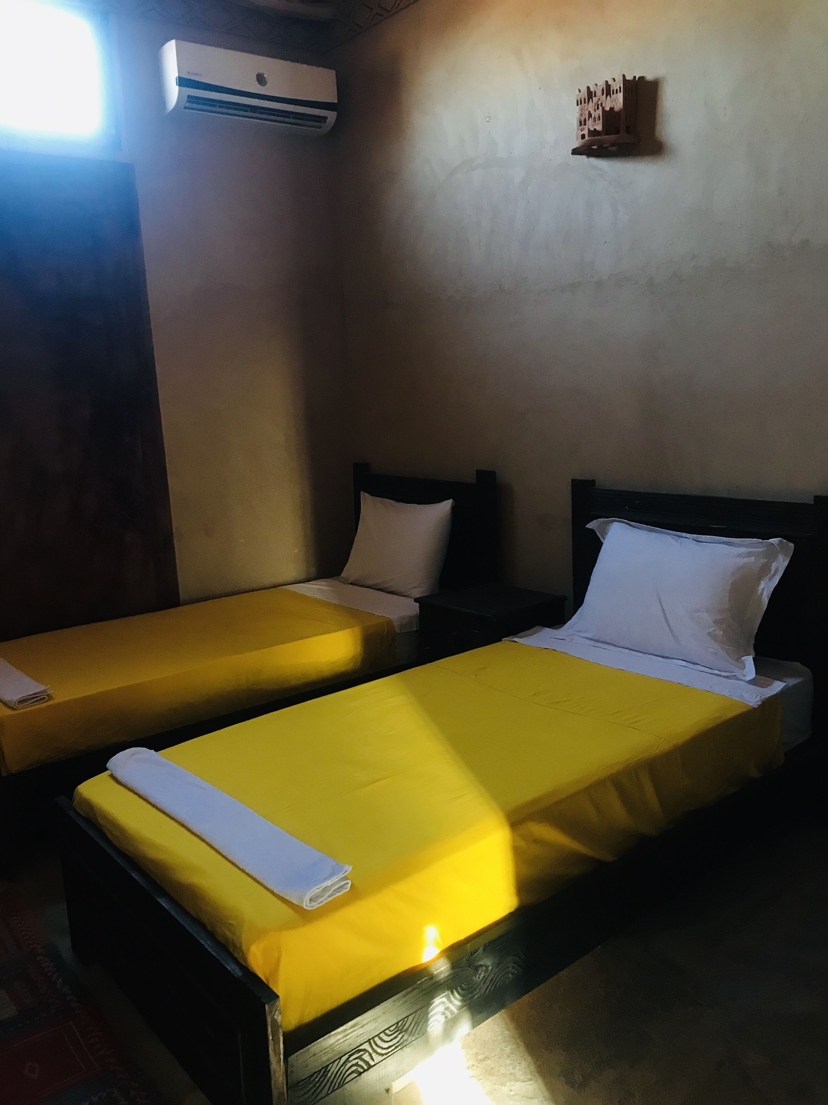 A room with two single beds, each with a yellow bedspread and white pillows. There is an air conditioning unit mounted on the wall above the beds.