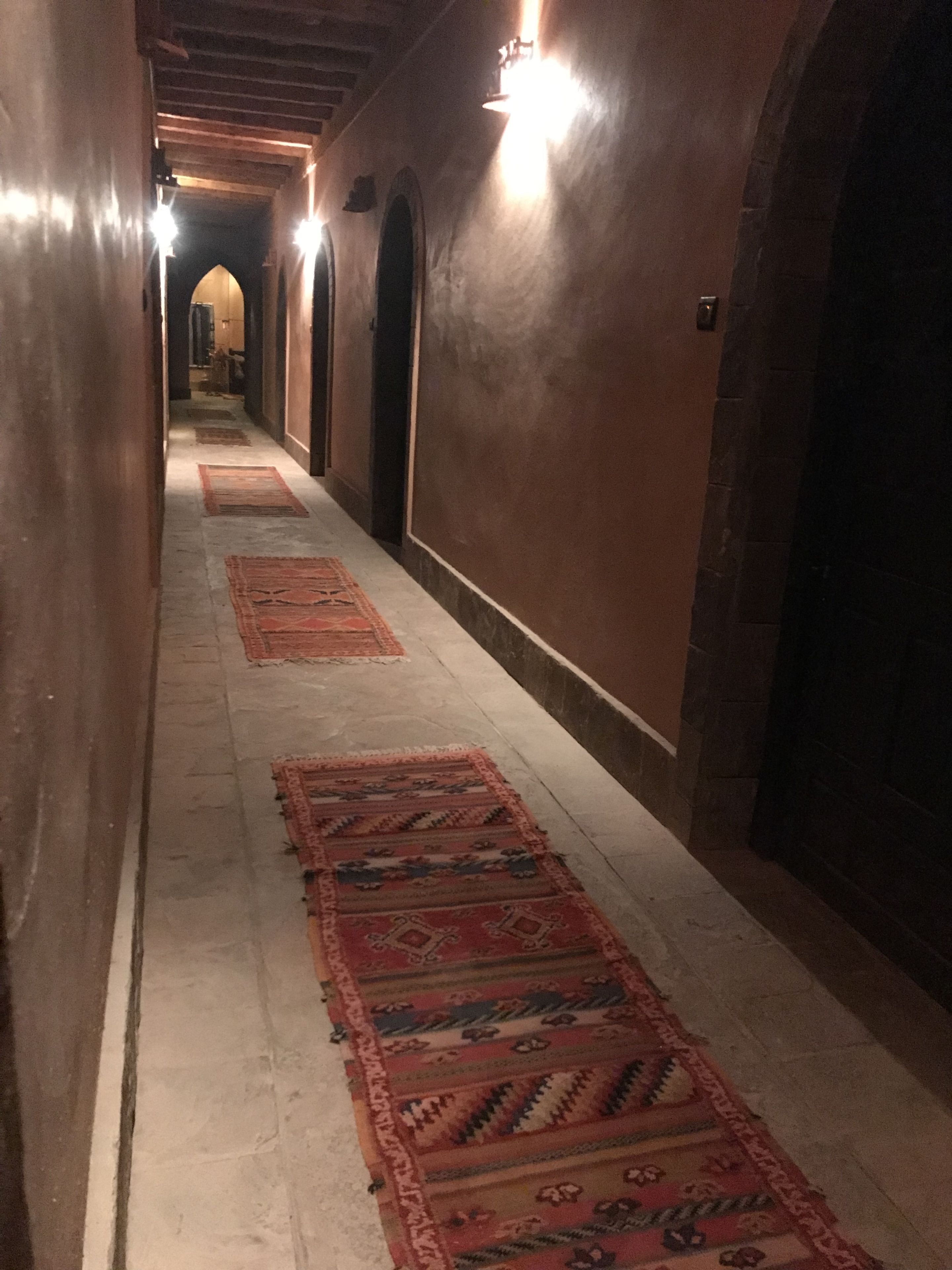 A corridor with patterned rugs, wooden doors, and wall-mounted lights.