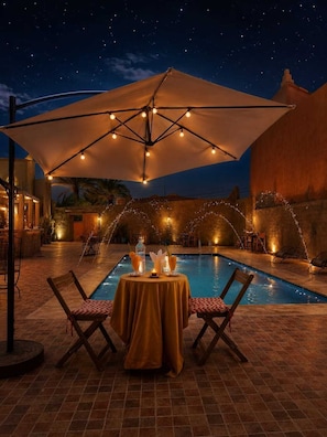 An outdoor pool area with a table and chairs under a canopy, surrounded by string lights. - Kasbah Ennakb (Nekob)