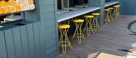 A covered outdoor seating area with wooden tables and yellow stools, a blue building with a window, and a stack of yellow towels on a shelf.