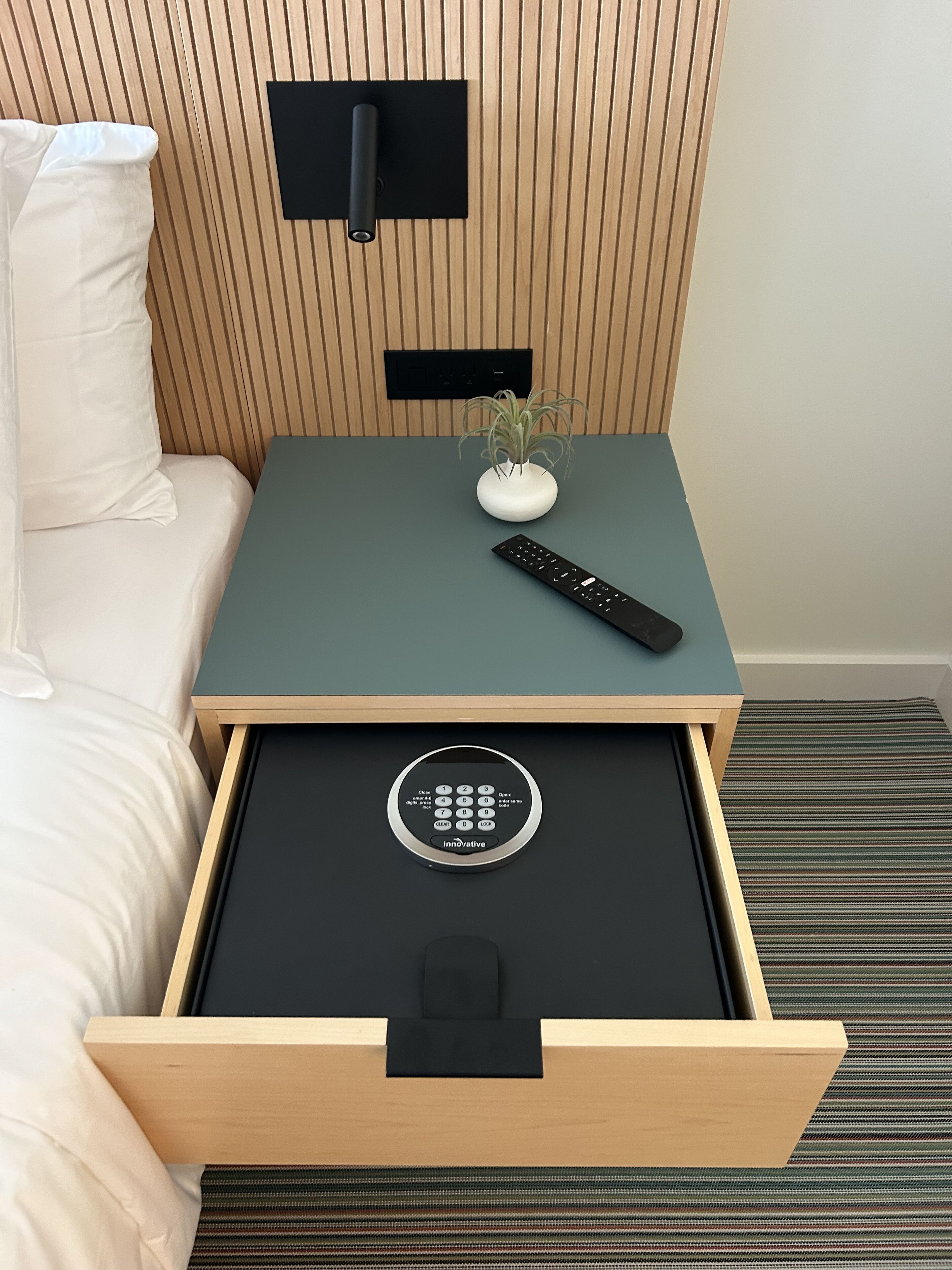 Ett hotellrum med en säng, ett litet bord med en fjärrkontroll, en krukväxt och en låda med en kontrollpanel.