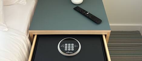 A hotel room with a bed, a small table with a remote control, a potted plant, and a drawer with a control panel.