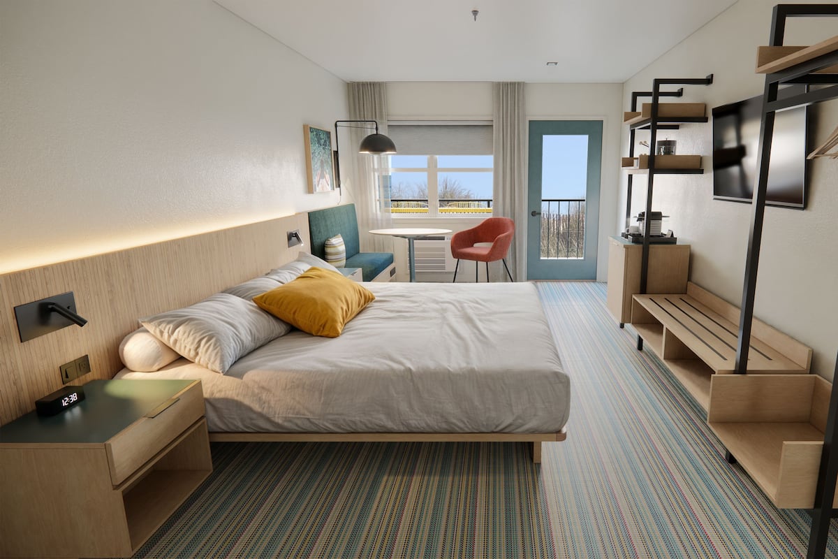 Una habitación de hotel moderna con una cama grande, un escritorio, una silla, una mesita, un banco, un perchero y una ventana con vistas.