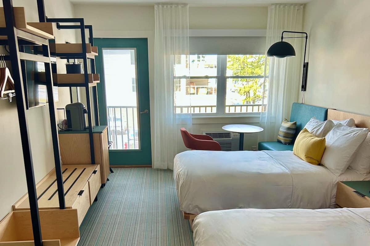 Habitación de hotel con dos camas, un escritorio, una silla, una mesita y una ventana con cortinas.