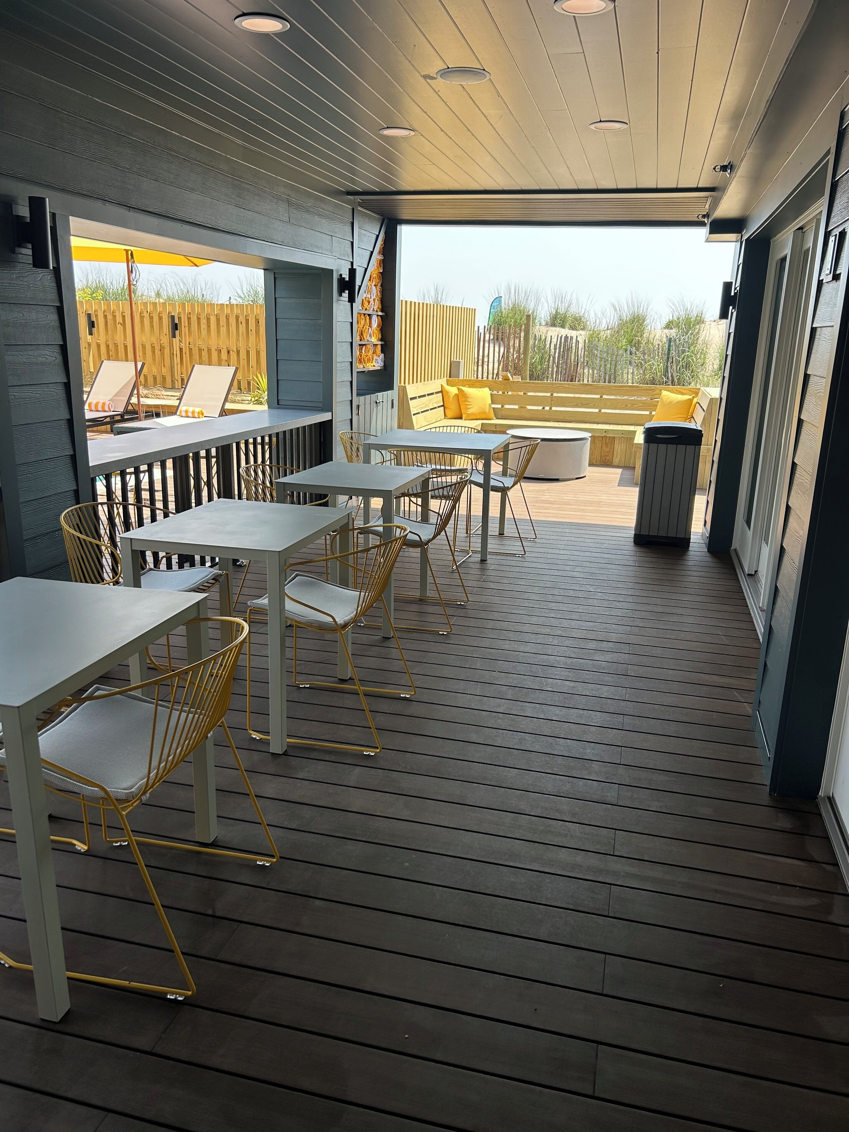An outdoor patio with wooden flooring, white tables, and yellow chairs.