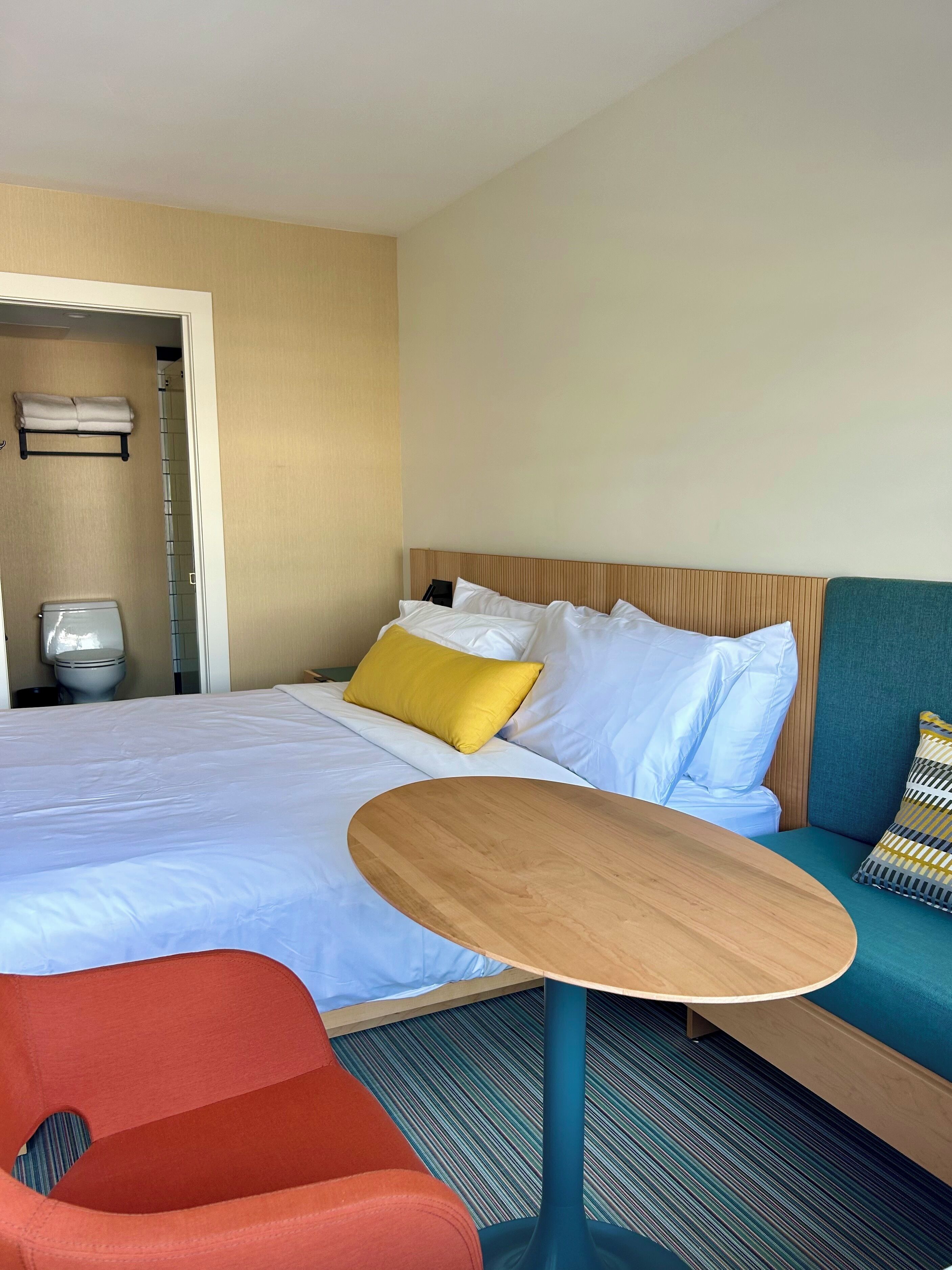 Habitación de hotel con una cama, una mesita de noche de madera, una silla roja, un sofá azul y un baño visible a través de una puerta abierta.