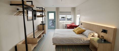 A modern hotel room with a bed, a desk, a chair, a small table, and a view of the city through the window.
