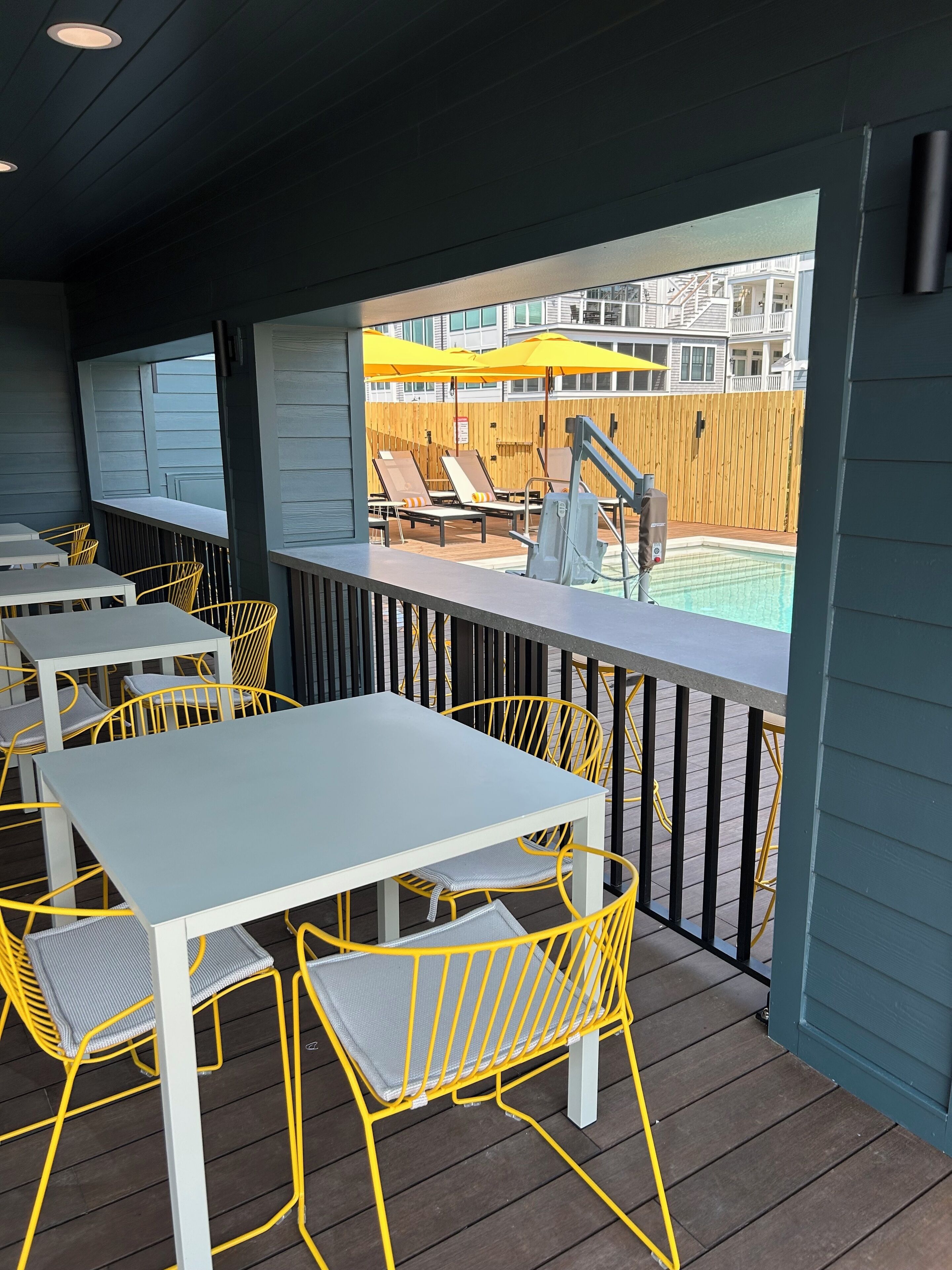 A covered outdoor seating area with tables and yellow chairs, overlooking a pool and wooden deck.