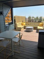An outdoor patio with a wooden table and chairs, a sofa with yellow cushions, and a round coffee table.