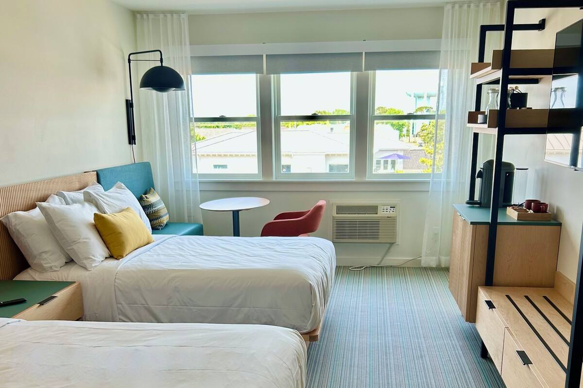 Habitación de hotel con dos camas, una mesita, una silla, un escritorio y ventana con vistas al exterior.