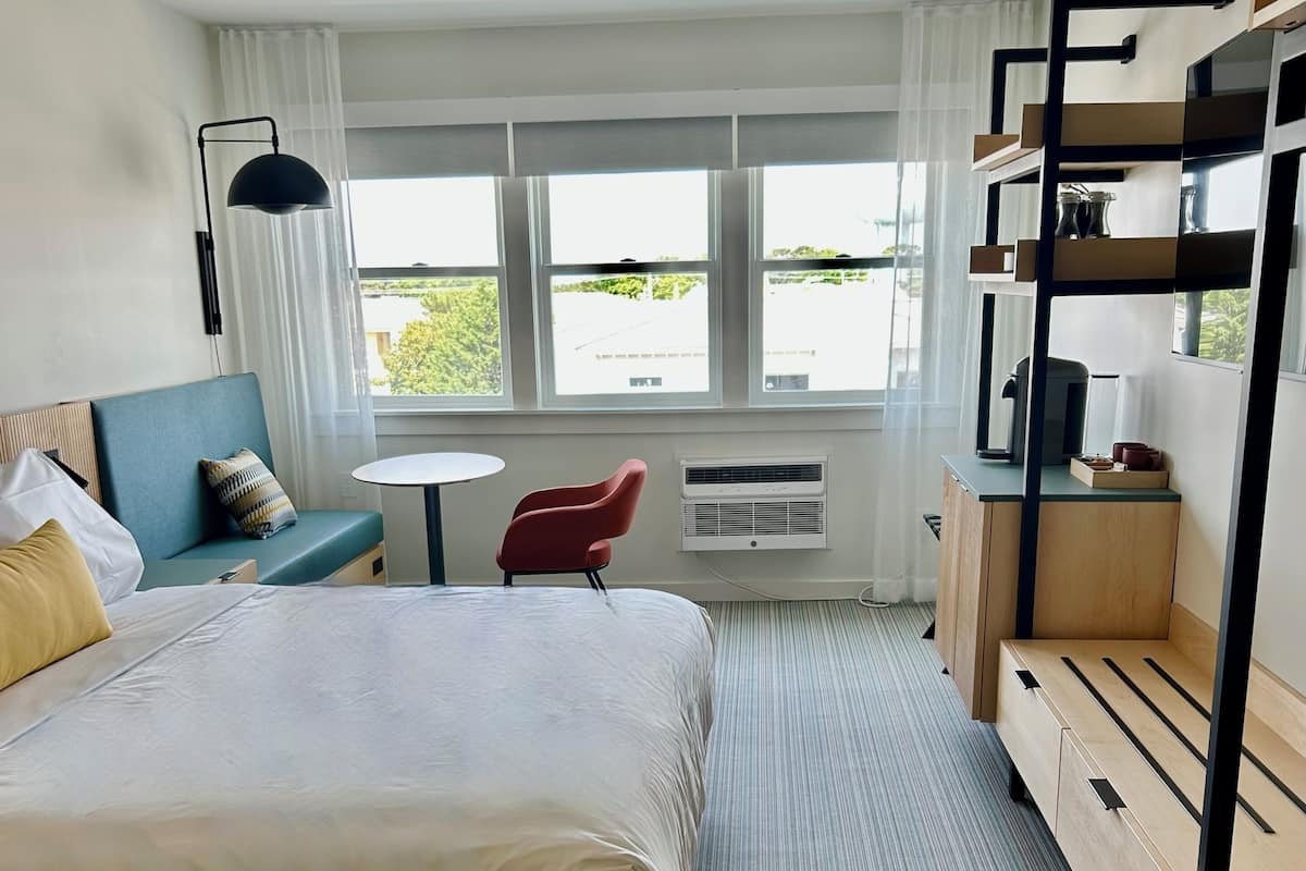 Habitación de hotel con una cama grande, una mesita pequeña, una silla roja, una cama litera y vistas al exterior a través de amplias ventanas.