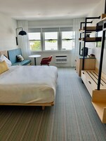 Une chambre d’hôtel avec un grand lit, une petite table, une chaise rouge, un lit superposé et une vue sur l’extérieur grâce à de grandes fenêtres.