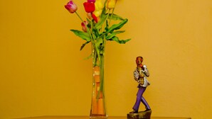 Een vaas met bloemen en een beeldje op een plank tegen een gele muur.