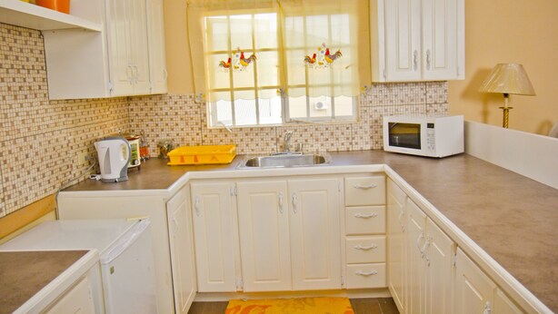 Een keuken met witte kastjes, een magnetron, een spoelbak en een raam met decoratieve gordijnen.