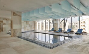 A modern indoor pool with clear glass walls, blue lounge chairs, and a view of surrounding buildings.