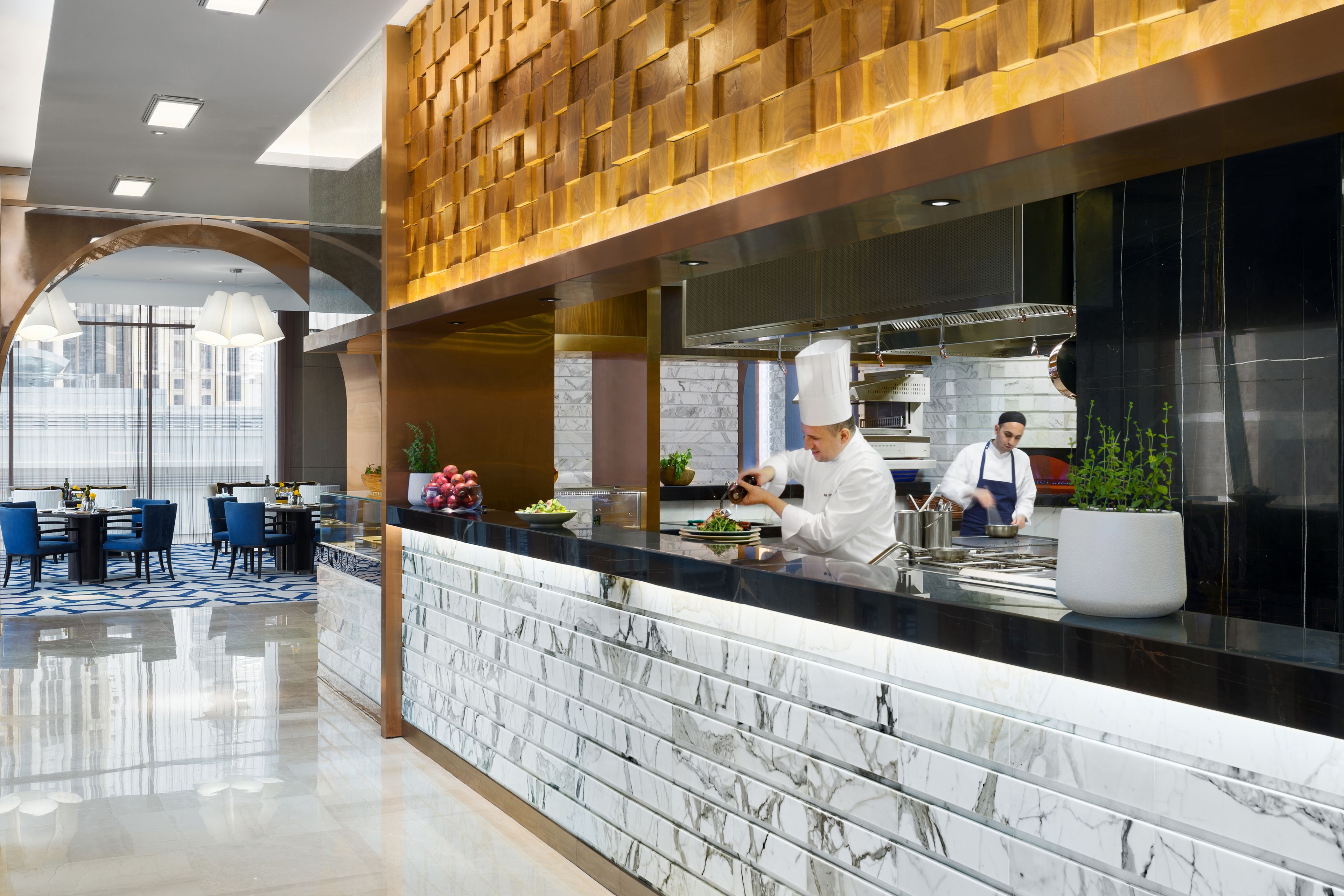 A modern restaurant with marble countertops, a chef preparing food, and a seating area with blue chairs.