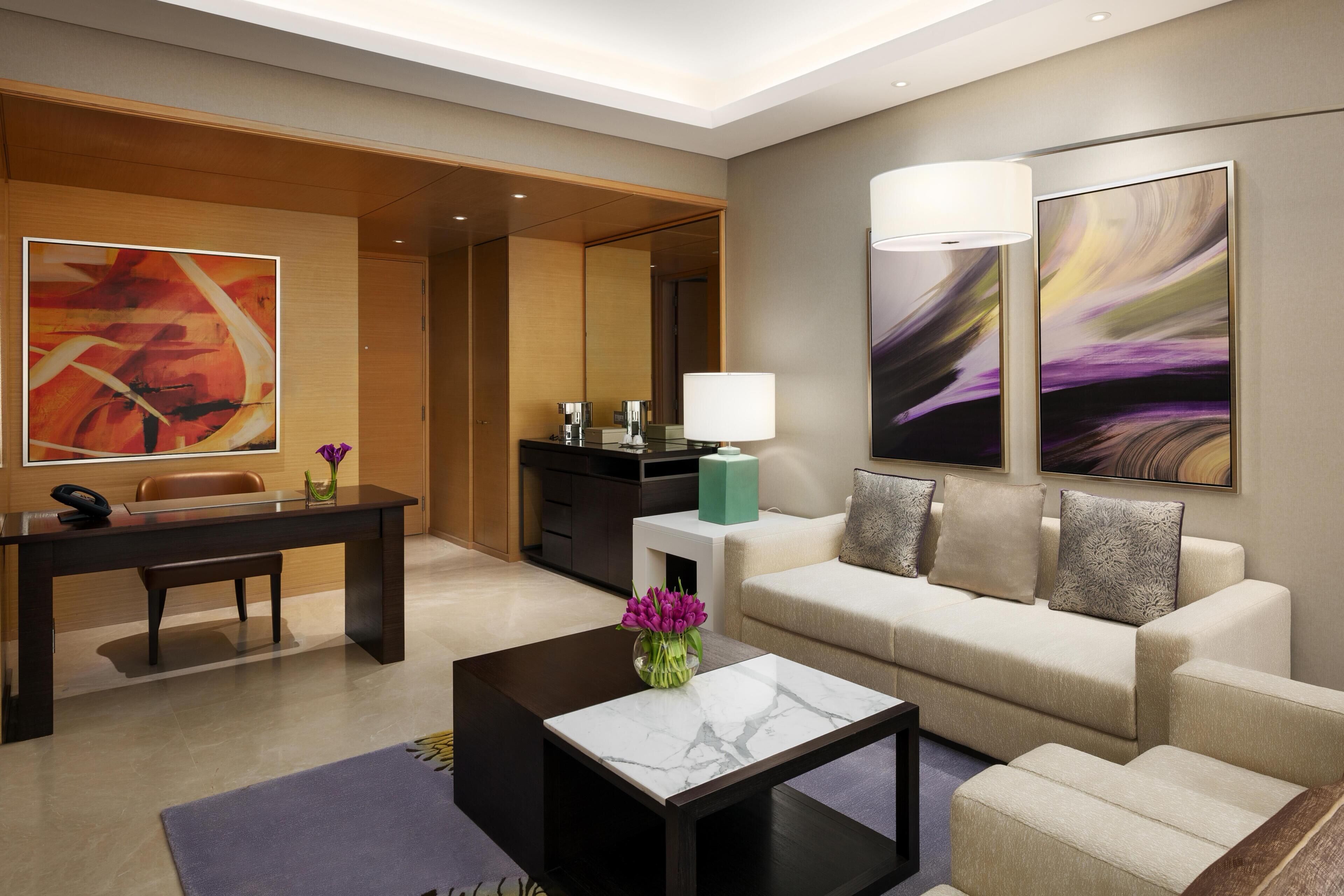 A modern hotel room with a sofa, a coffee table, a desk, and a lamp.