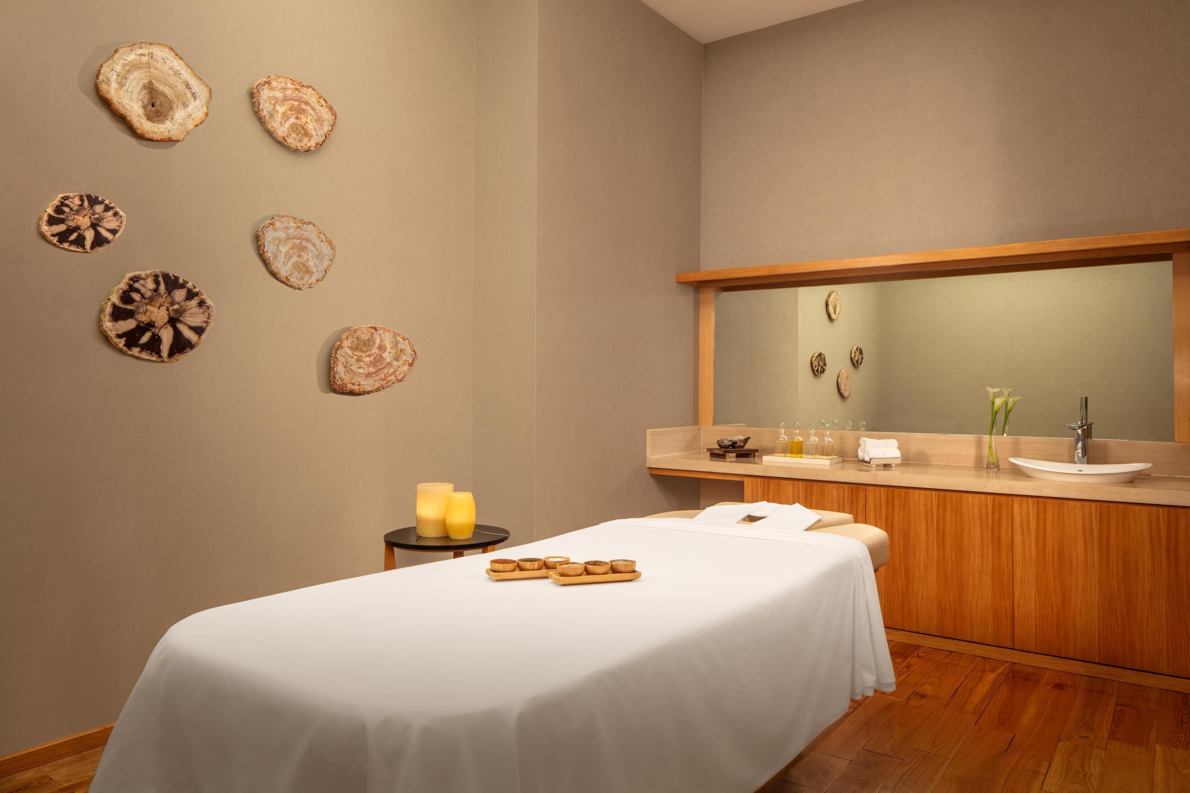 Une salle de spa équipée d’une table de massage, de bougies, d’une petite table en bois et d’un grand miroir placé au-dessus d’un lavabo.