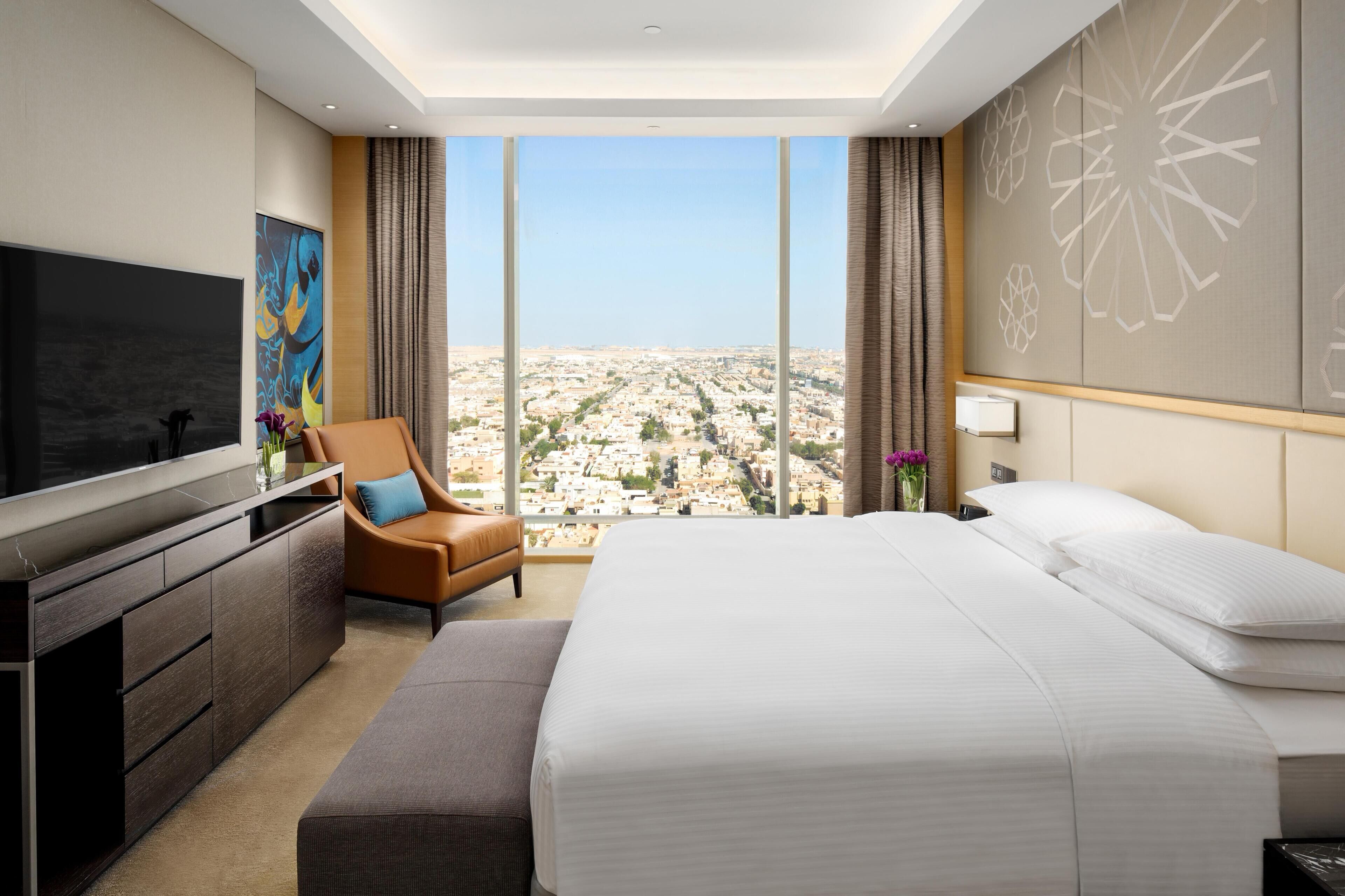 A modern hotel room with a large bed, a TV, a seating area, and a city view.