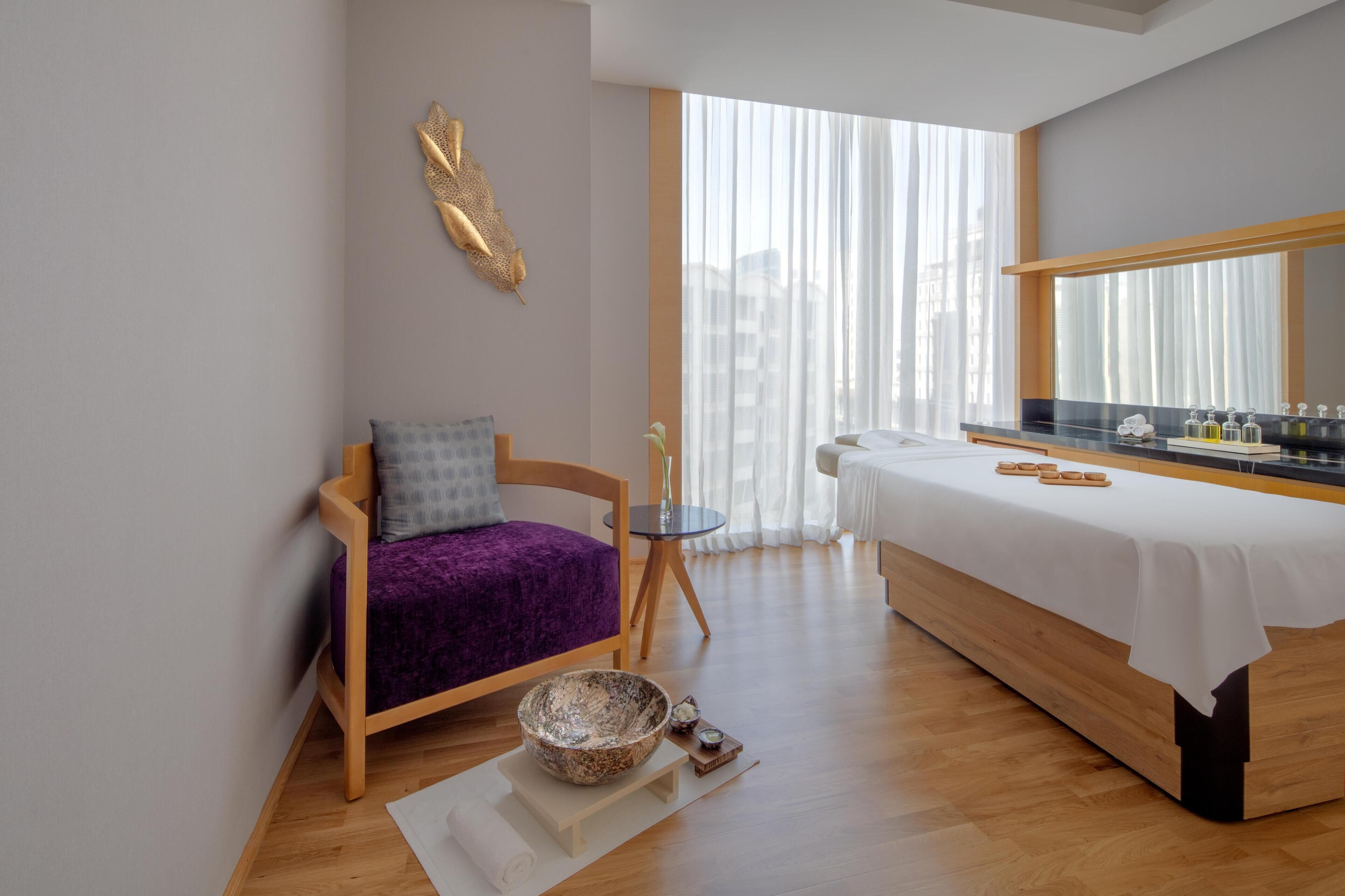 A modern room with a wooden bed, a small table with a bowl, a chair with a purple cushion, and a large window with sheer curtains.