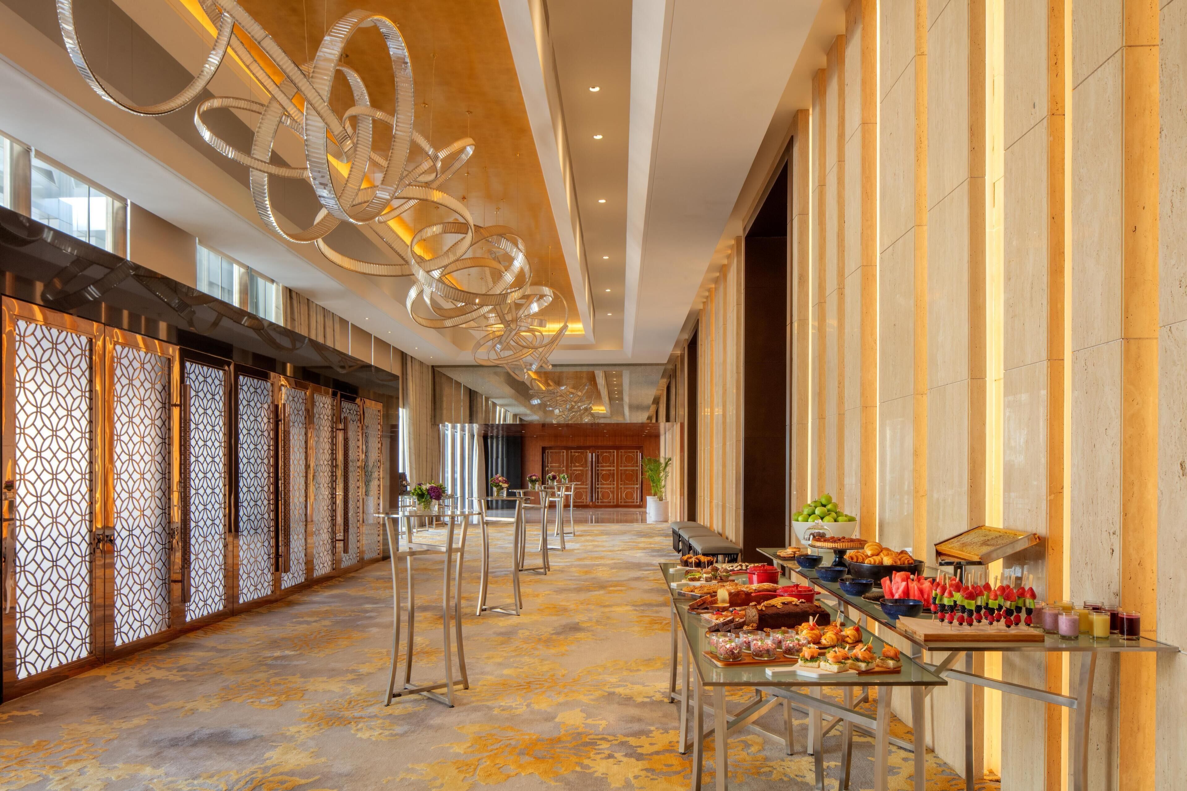 A spacious corridor with a long table displaying food and drinks, a modern chandelier, and a decorative wall with vertical lines.