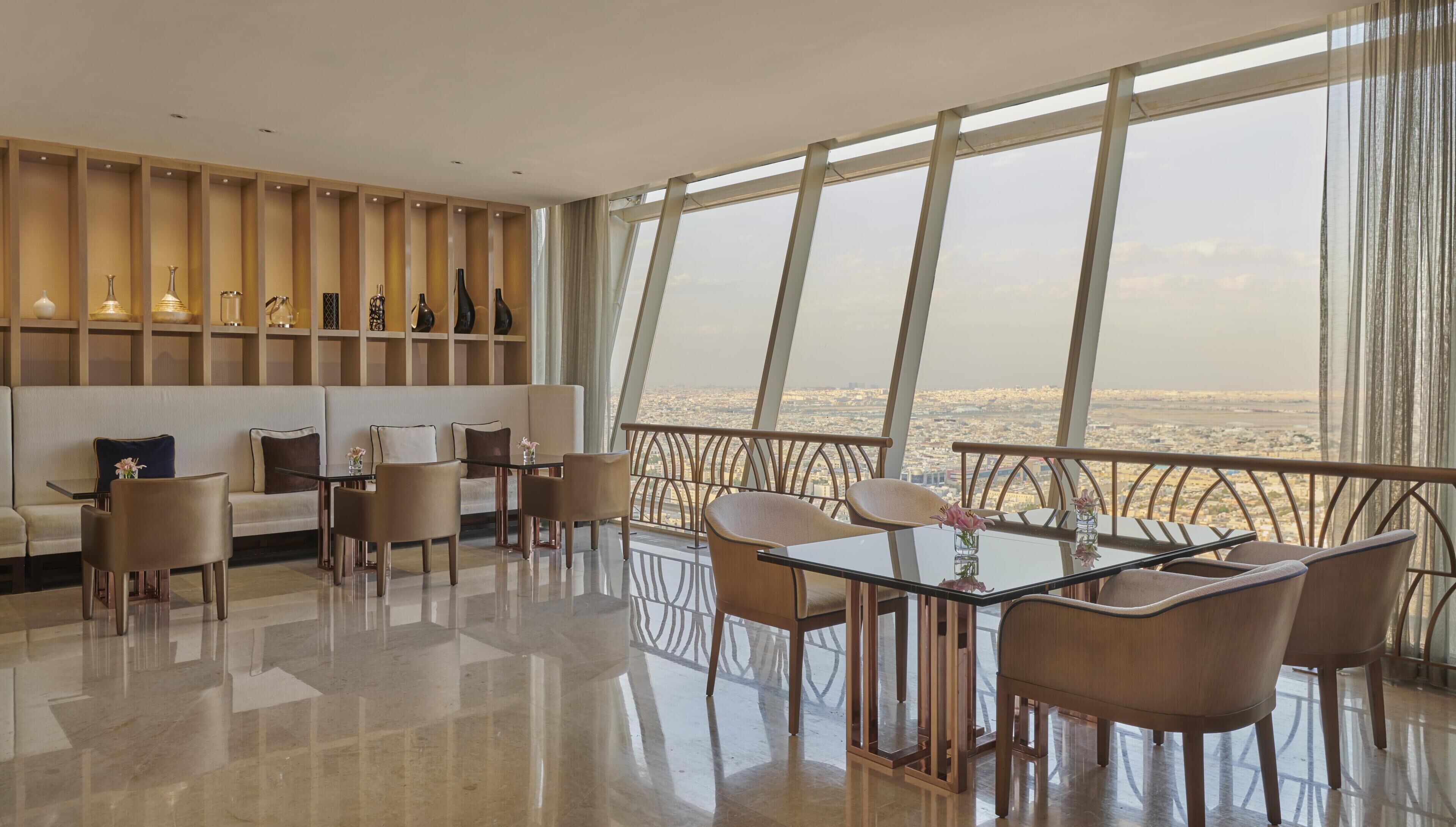 Uma sala de estar moderna com sofá, cadeiras, mesa de vidro e vista da cidade através de grandes janelas.
