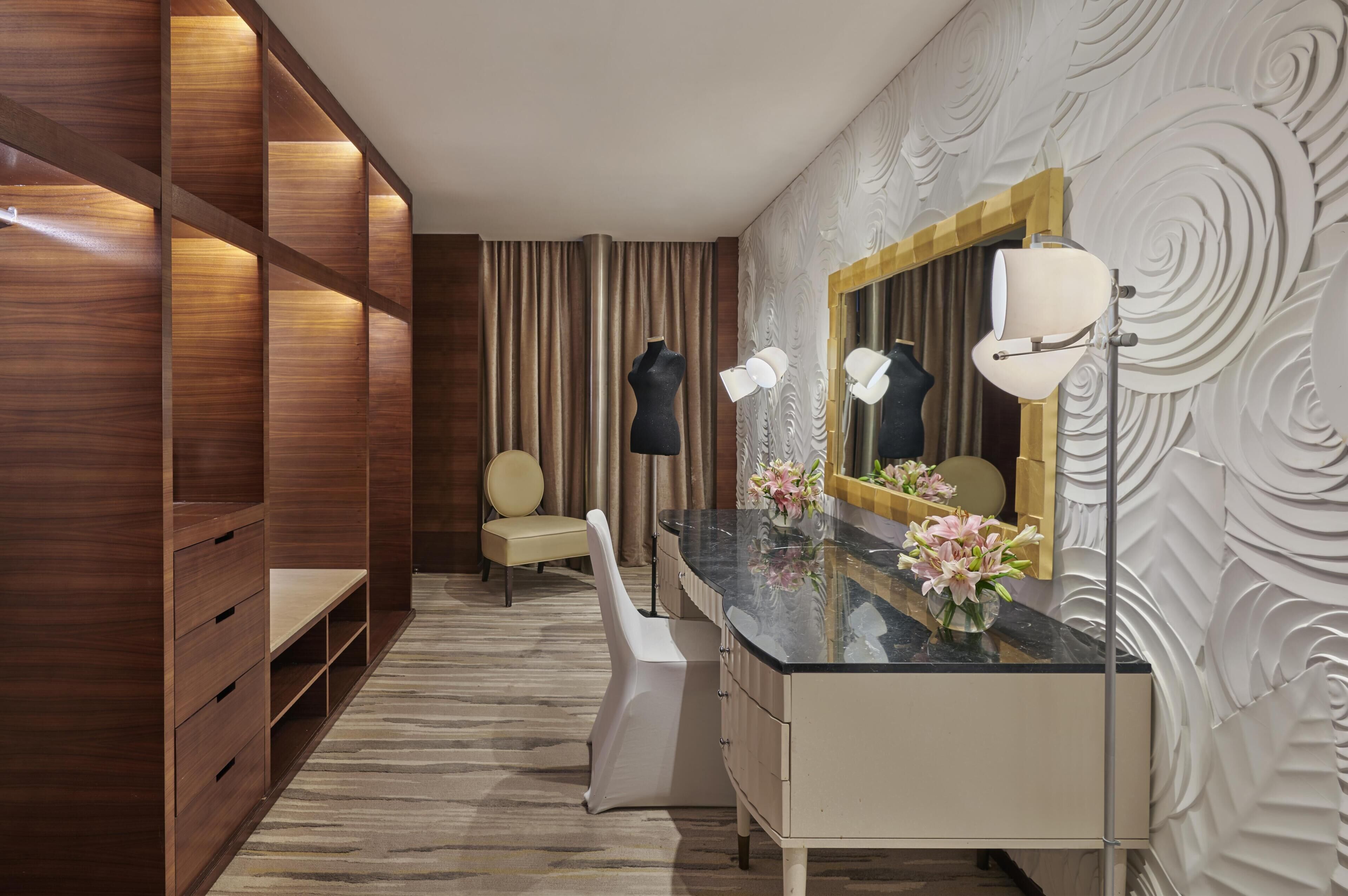 A hotel room with a vanity, a mirror, a chair, a wardrobe, and a patterned wall.