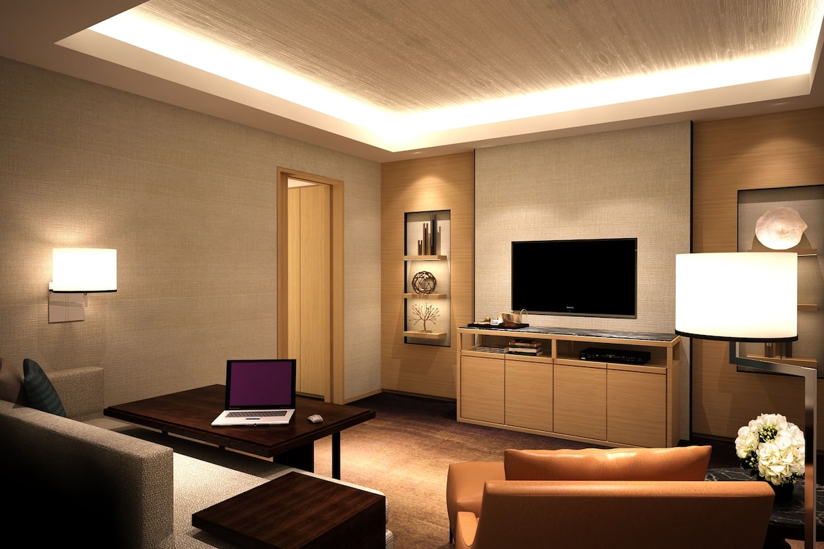 A modern hotel room with a flat-screen TV, a laptop on a wooden table, a sofa, and a TV stand with a flat-screen TV and a lamp.