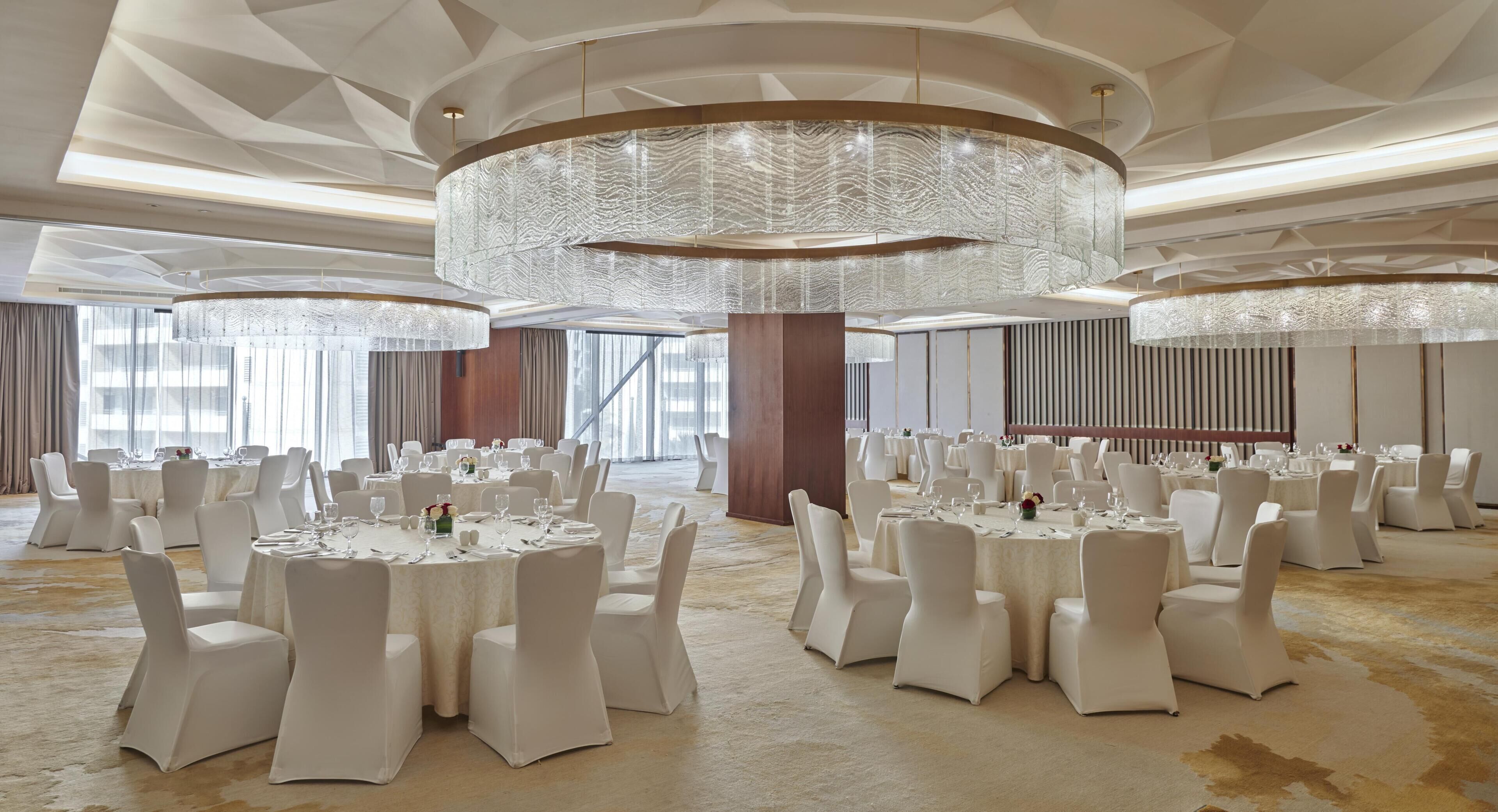 A spacious banquet hall with round tables, white chairs, and large chandeliers.