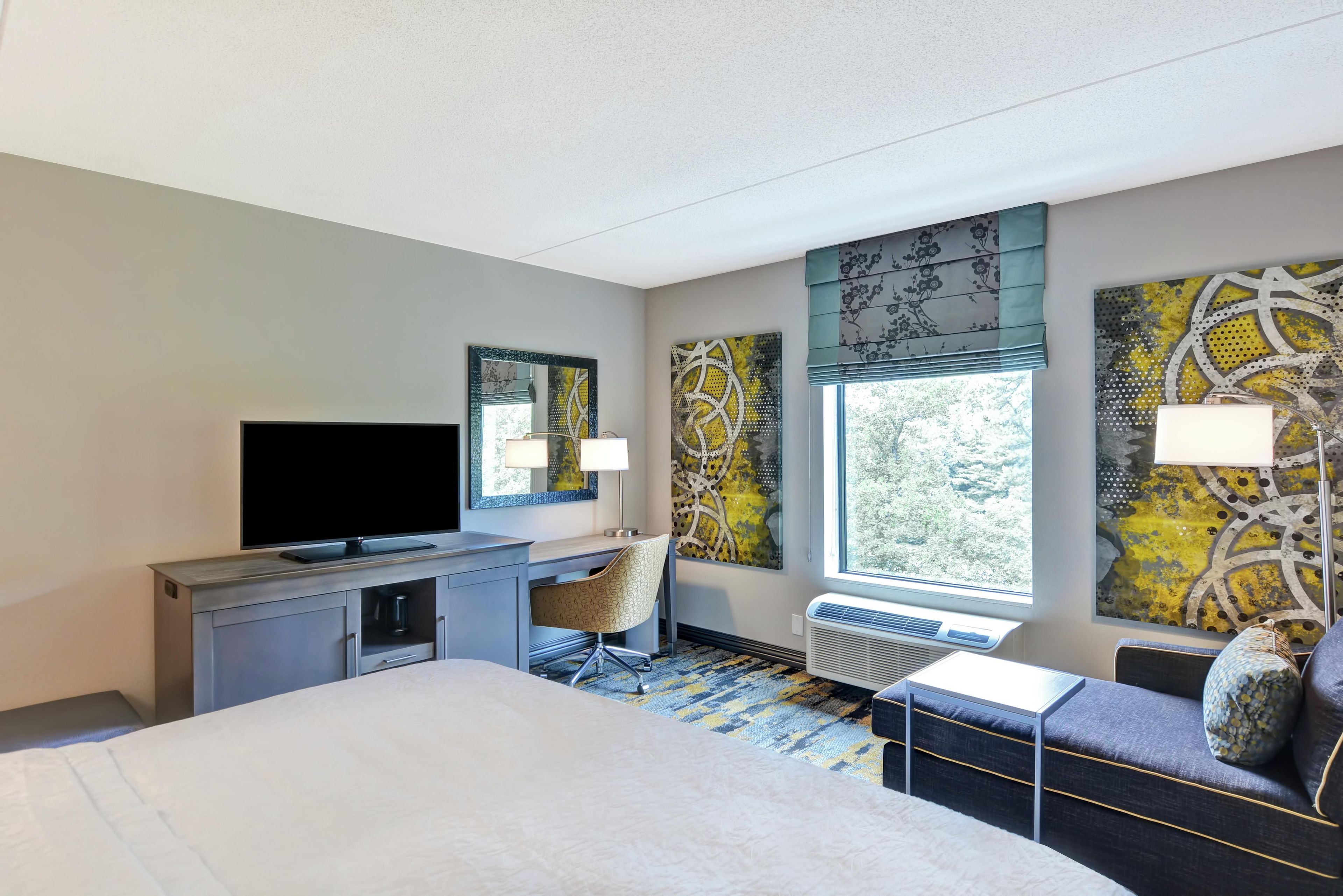 A hotel room with a bed, desk, chair, television, artwork, and a window.