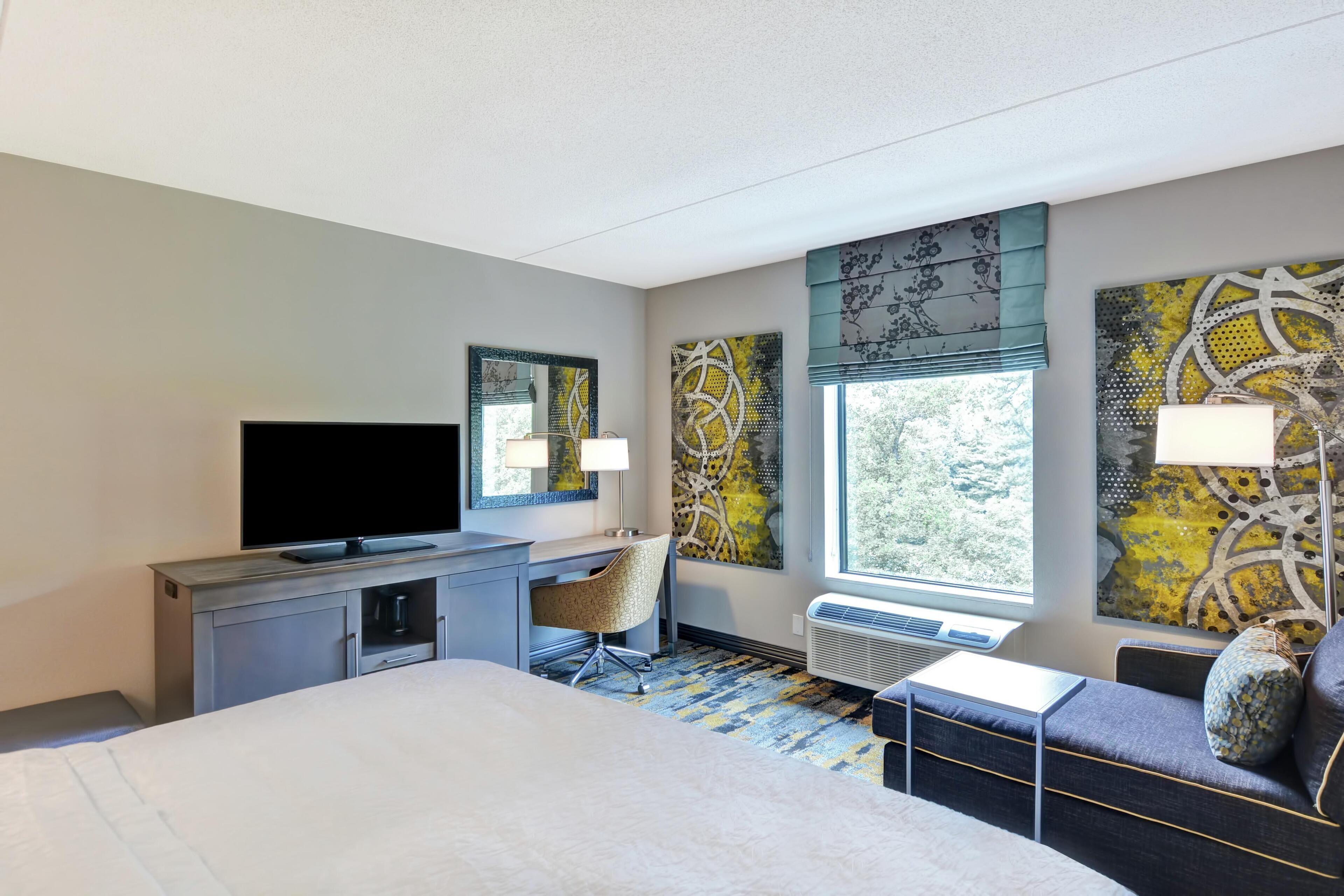 A hotel room with a bed, desk, chair, television, artwork, and a window.