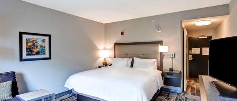 A hotel room with a bed, bedside tables, a TV, a painting on the wall, and a door leading to another room.