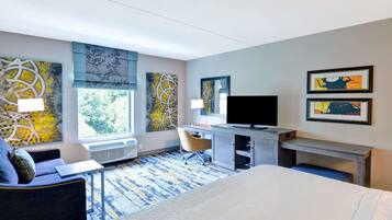 Una habitación de hotel con cama, un escritorio con computadora, una silla, un televisor y obras de arte en las paredes.
