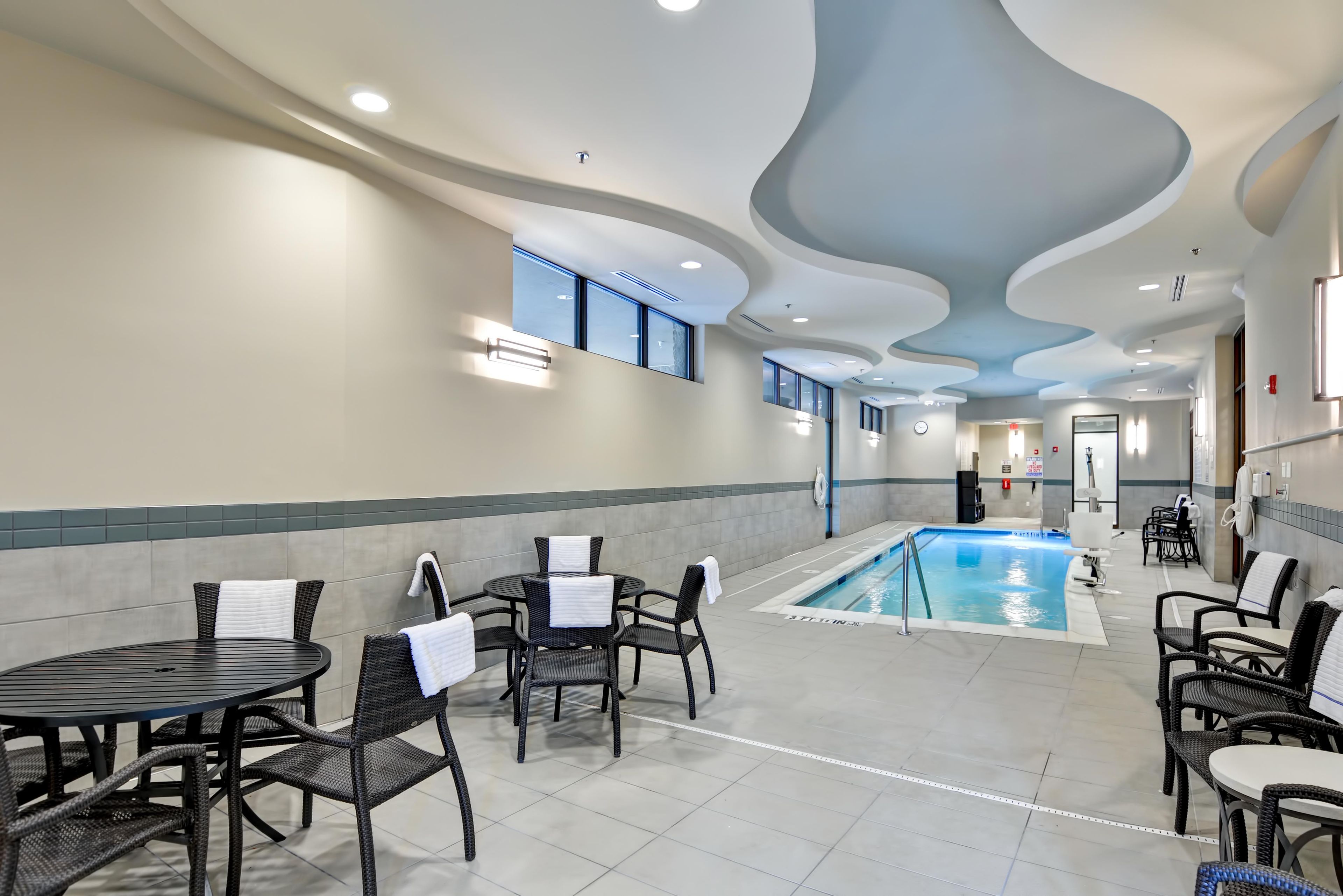 A pool area with lounge chairs and tables, a pool with a blue interior, and a modern ceiling design.