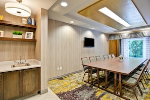 Una sala de reuniones moderna con una larga mesa, sillas, un televisor de pantalla plana y un ventanal con cortinas.