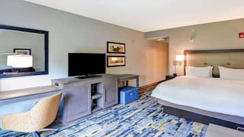 A hotel room with a bed, a desk with a television, a chair, and a blue suitcase.