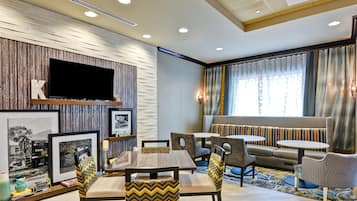 Habitación de hotel con televisor, cuadros enmarcados, sofá y zona de comedor con sillas y mesas.
