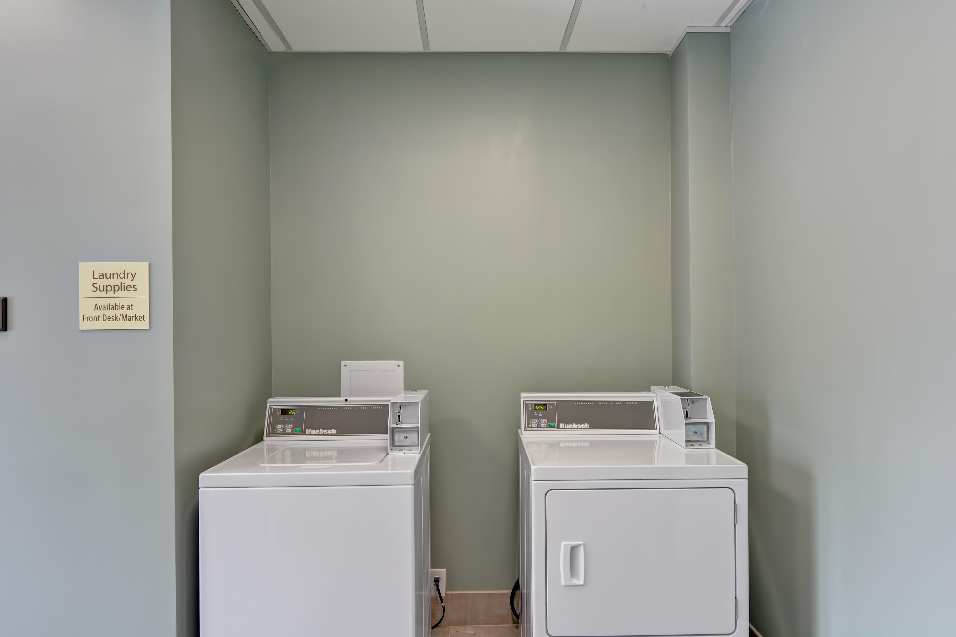 Une buanderie équipée de deux machines à laver à chargement frontal et d’un sèche-linge, un panneau indiquant que le nécessaire pour faire la lessive est disponible à la réception/à l’épicerie, et un mur de couleur vert clair.