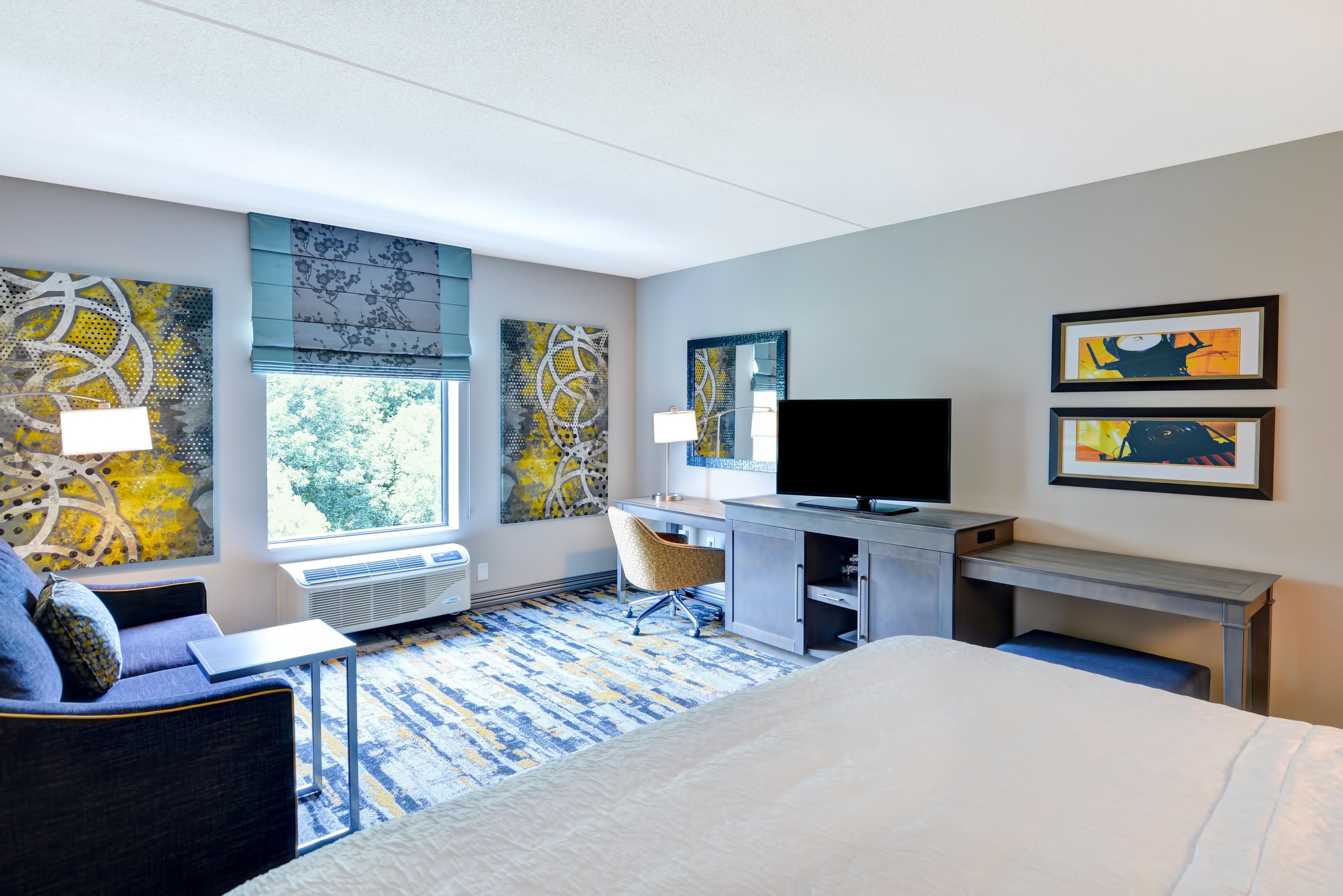 A hotel room with a bed, a desk with a flat-screen TV, a chair, and artwork on the walls.