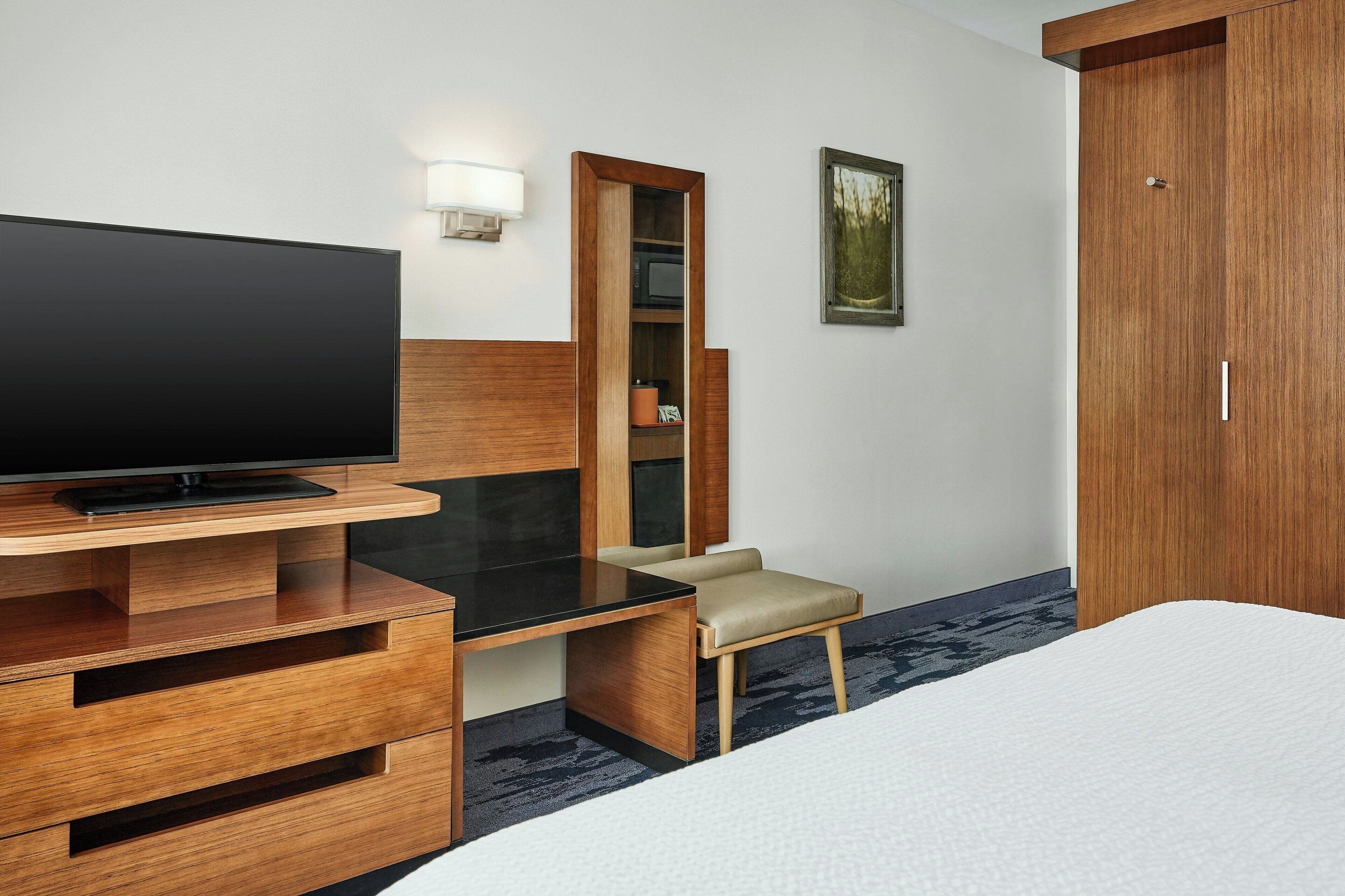 A hotel room with a flat-screen TV on a wooden desk, a small chair, a bed with white bedding, a wooden wardrobe, and a framed picture on the wall.