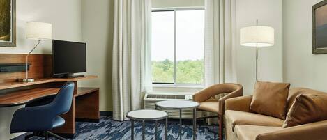 Ein Hotelzimmer mit Schreibtisch, blauem Bürostuhl, braunem Sofa, zwei kleinen runden Tischen und einem großen Fenster mit Vorhängen.