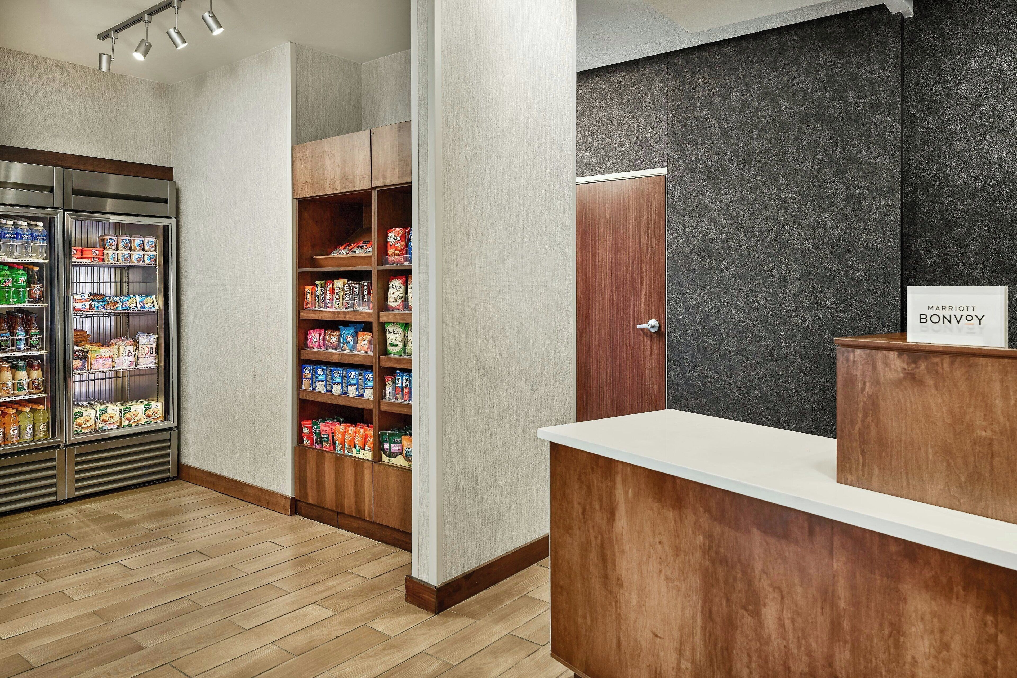 A hotel reception area with a wooden reception desk, a refrigerator stocked with beverages, and a pantry with various food items.
