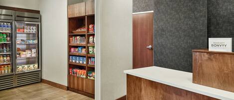 A hotel reception area with a wooden reception desk, a refrigerator stocked with beverages, and a pantry with various food items.