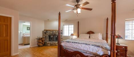 A spacious bedroom with a four-poster bed, a ceiling fan, a stone fireplace, and a view of the adjacent bathroom.