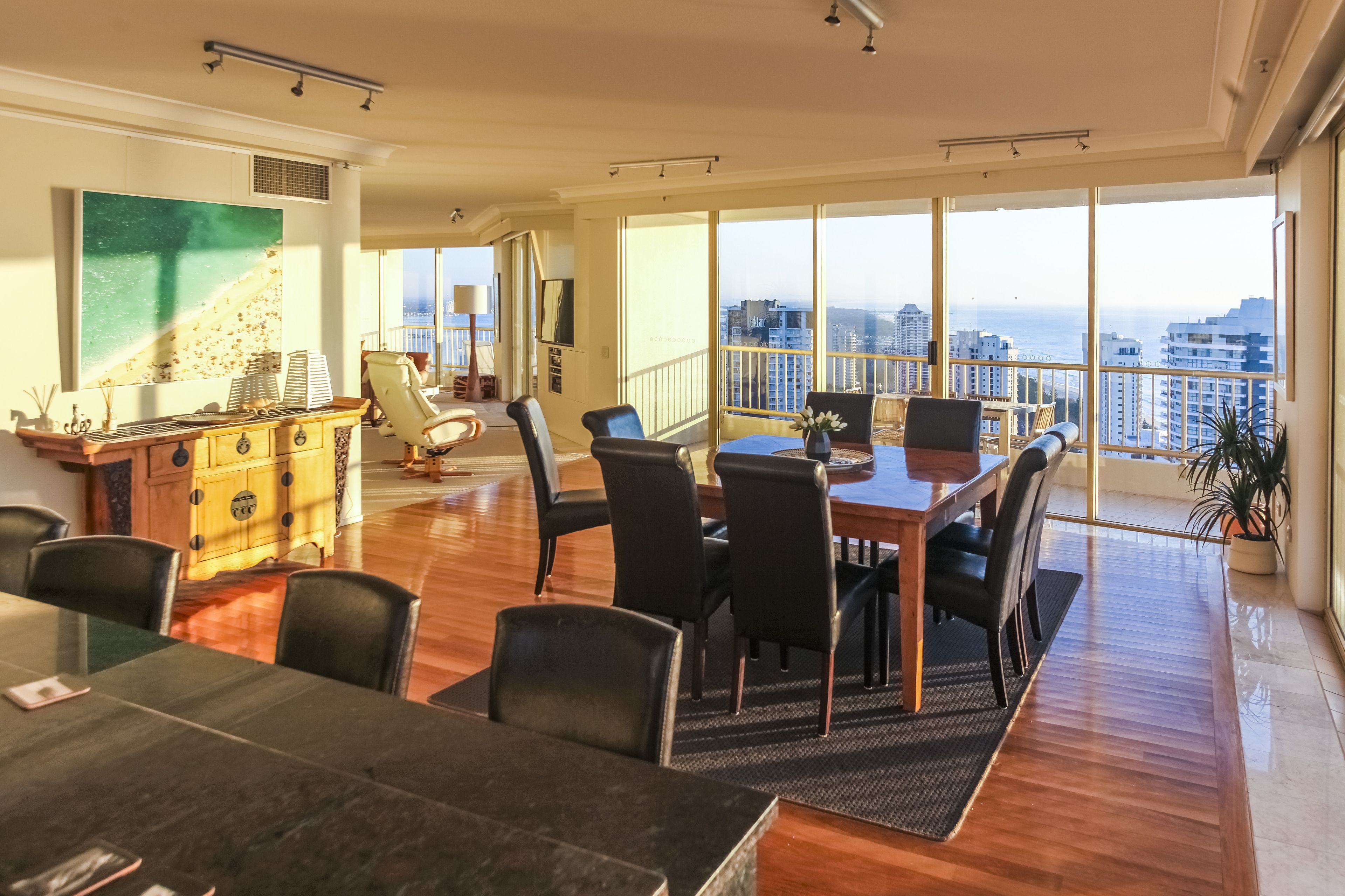 A spacious dining area with a high ceiling, large windows offering a city view, and a modern kitchen with a wooden cabinet.